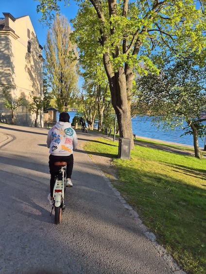 A person riding a bicycle on a paved path alongside a body of water, with trees and a building in the background. 3