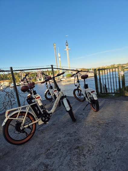 Three electric bicycles parked near a waterfront with a fence and a view of a cityscape in the background. 5
