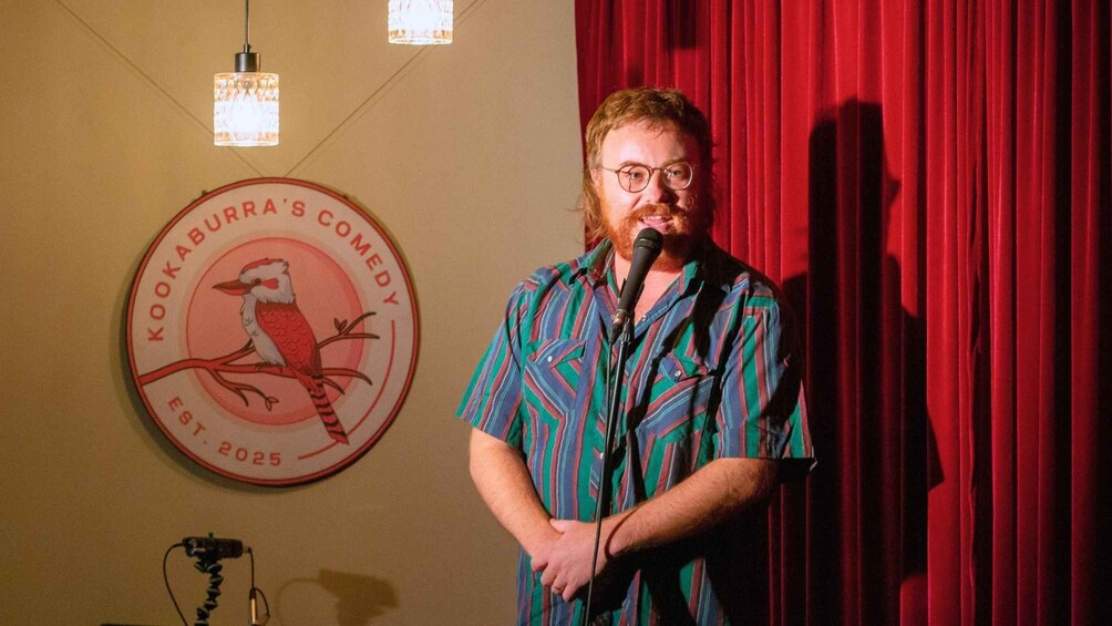 A man standing in front of a microphone with a red curtain backdrop and a sign that reads 'Kookaburra's Comedy' on the wall. 5