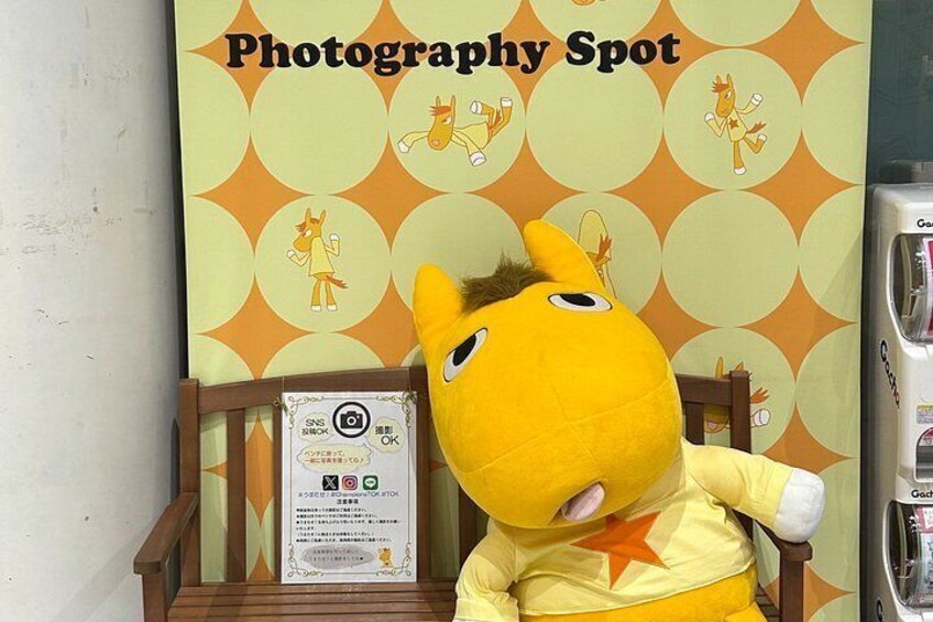 A yellow plush toy seated on a wooden chair with a 'Photography Spot' sign in the background. 2