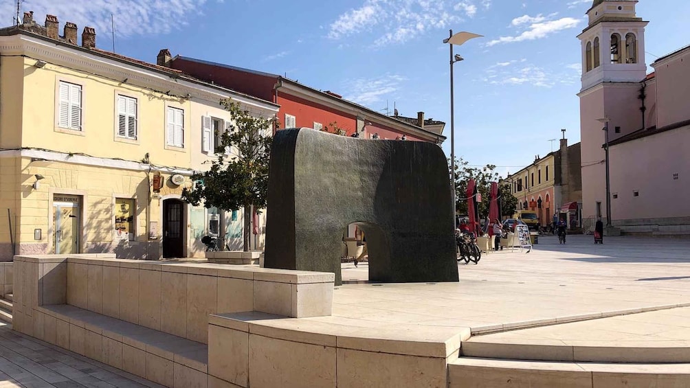 A public square with a large stone sculpture, buildings with classic architecture, and a church tower in the background. 2
