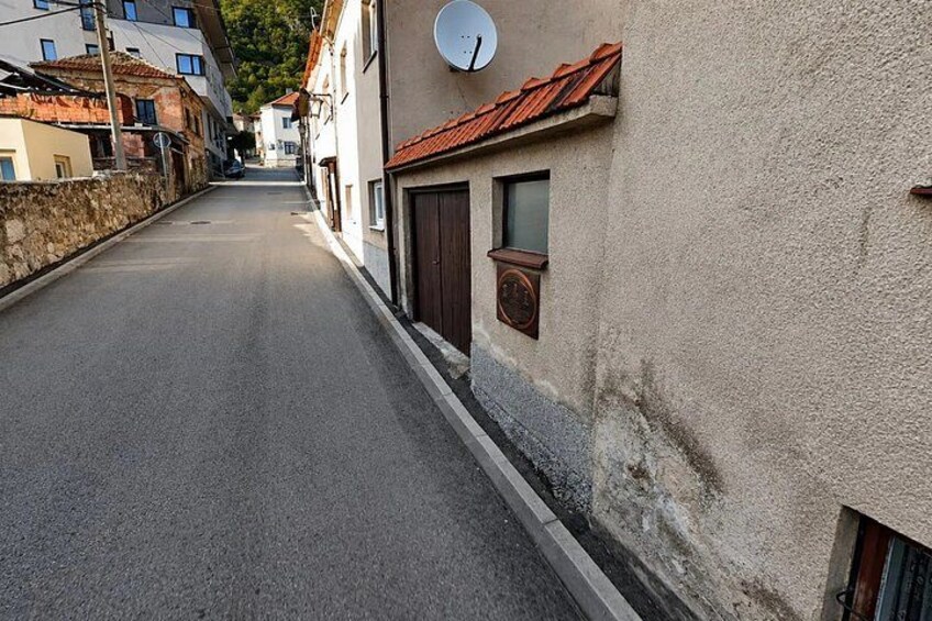 A narrow street with buildings on either side, a satellite dish on one building, and a stone wall on the left. 5