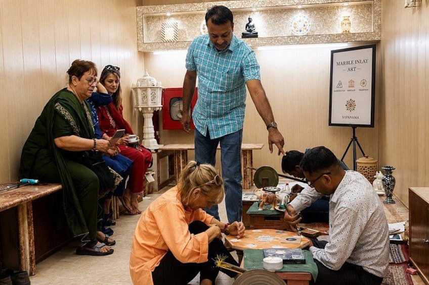A group of people engaged in a craft activity, with a framed artwork and a lamp in the background. 1