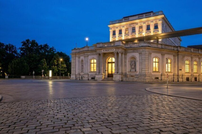 A historic building with 'ALBERTINA' signage, a cobblestone plaza, and a clear evening sky. 5