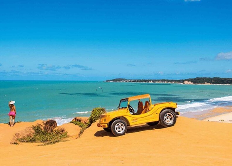 A yellow off-road vehicle on a sandy beach with clear blue water and a person in a pink swimsuit. 5