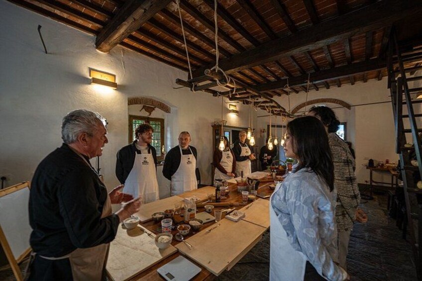 A group of people in a rustic kitchen with wooden beams, hanging lights, and a long table with cooking utensils and ingredients. 1