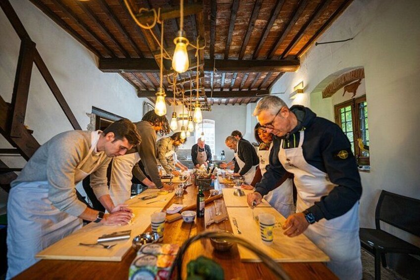A group of people in aprons preparing food at a long wooden table in a rustic kitchen with exposed wooden beams and hanging light bulbs. 3