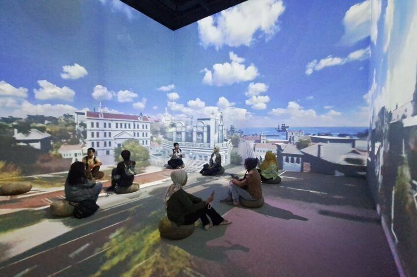 A group of people sitting on the ground, facing a large digital display of a cityscape. 2