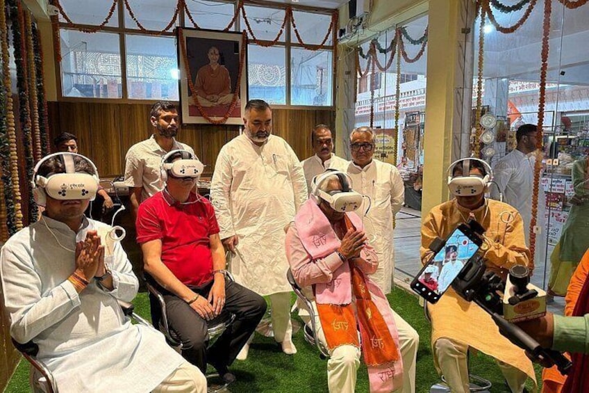 People wearing VR headsets in a room with a green carpet, a framed picture on the wall, and a person holding a camera. 4
