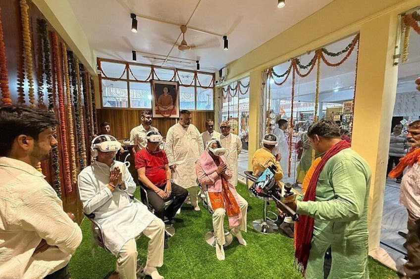 A group of people in traditional attire sitting on chairs, some wearing VR headsets, in a room with a green carpet and a Buddha statue on the wall. 5