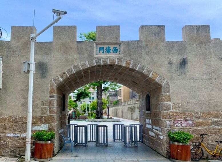 An arched stone gateway with a sign, potted plants, a bicycle, and a security camera. 2