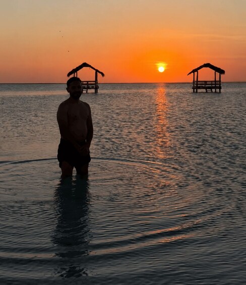A person standing in shallow water at sunset with two structures in the background. 1