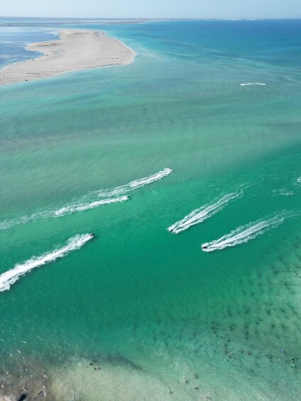 A coastal area with clear turquoise waters, a sandy beach, and a boat creating wake patterns. 4
