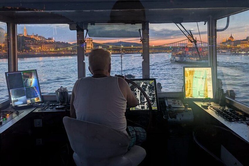 A man operating a ship's control panel with a cityscape and bridge in the background. 5