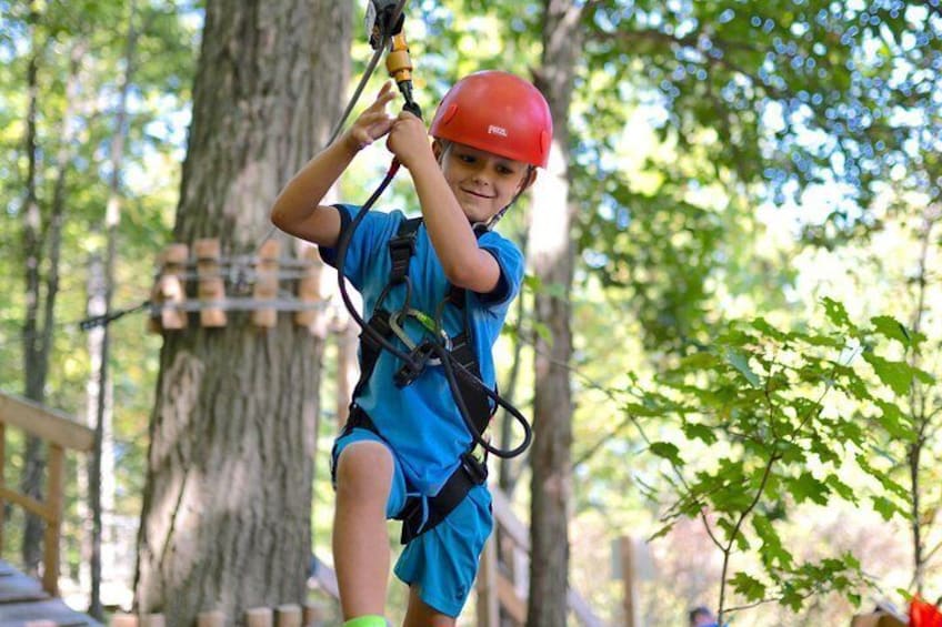 A person in a blue shirt and red helmet engaged in an outdoor activity, possibly zip-lining, with trees and a wooden structure in the background. 5