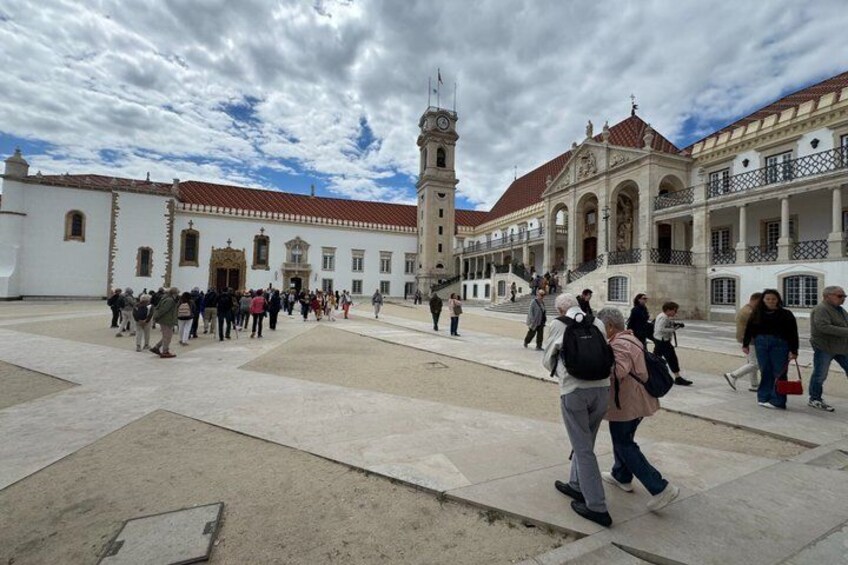 A historic plaza with a prominent clock tower, a large white building with arches, and a crowd of people. 4