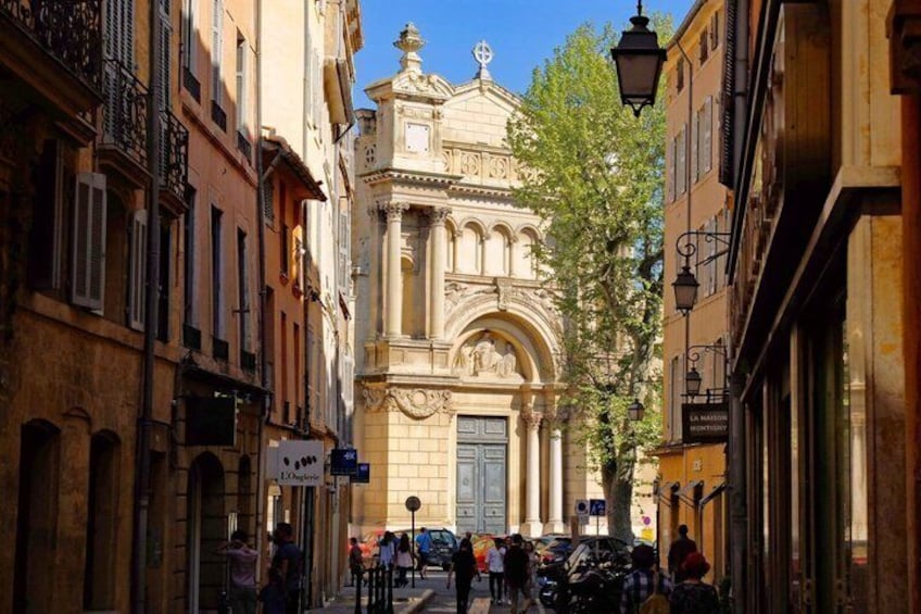 A narrow street with a historic church, pedestrians, and parked cars. 1