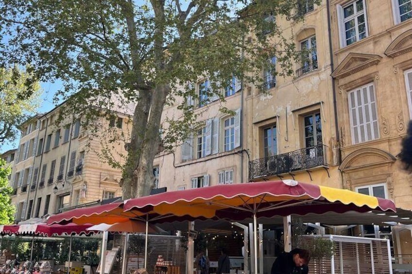 A street market with a red and yellow umbrella, a tree, and old buildings. 5
