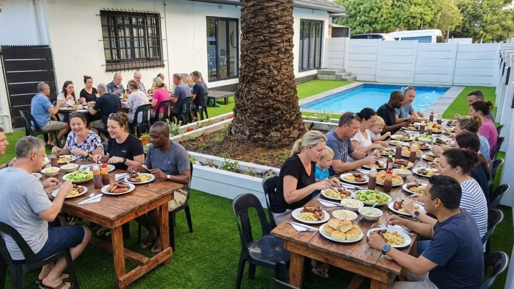 Un grupo de personas cenando al aire libre en una larga mesa de madera con una piscina al fondo. 4