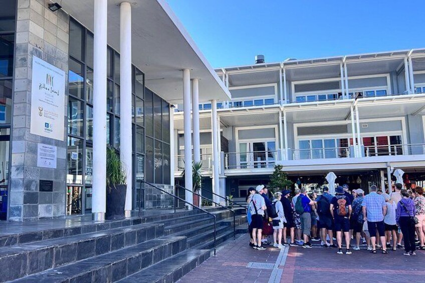 A modern building with a glass facade, a set of stairs leading up to a balcony, and a group of people gathered outside. 1