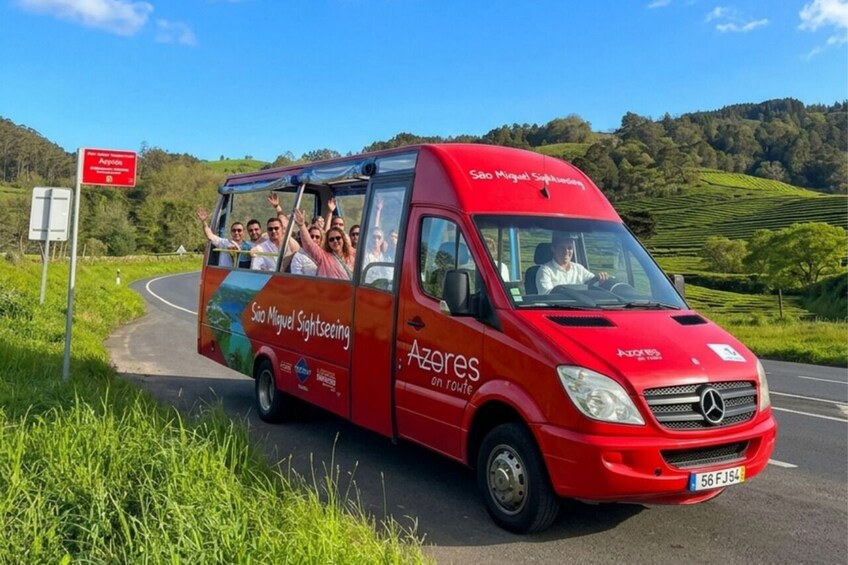En röd sightseeingbuss full med passagerare som vinkar, med texten ”São Miguel Sightseeing” på sidan, kör på en landsväg med gröna fält och kullar. 2