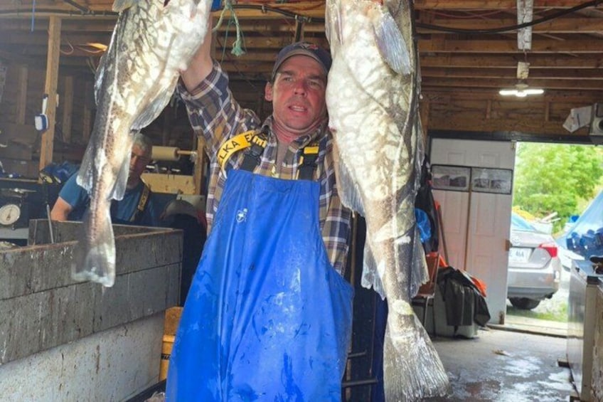 A person holding two fish, wearing a plaid shirt and blue overalls, in a garage with a car and tools visible. 3