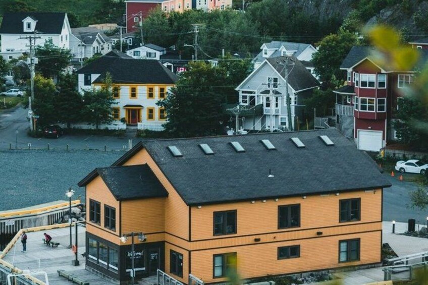A multi-story building with a sloped roof, a street with parked cars, and a pedestrian walkway. 3