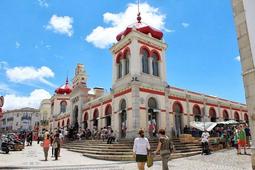 A historic building with red domes and a clock tower, surrounded by a cobblestone square with pedestrians. 1