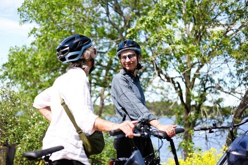 Two people on bicycles, one wearing a helmet, standing by a body of water with trees in the background. 3