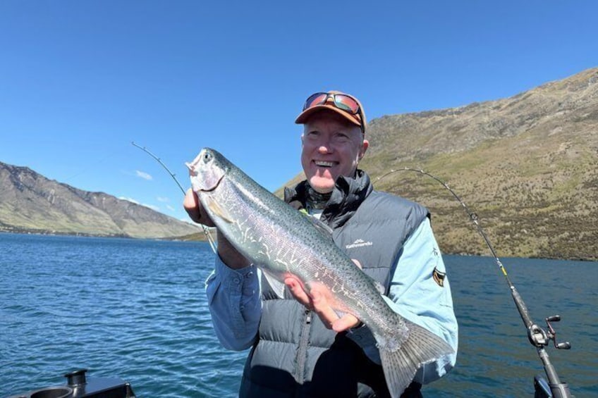 A person holding a large fish on a boat with a lake and mountains in the background. 1