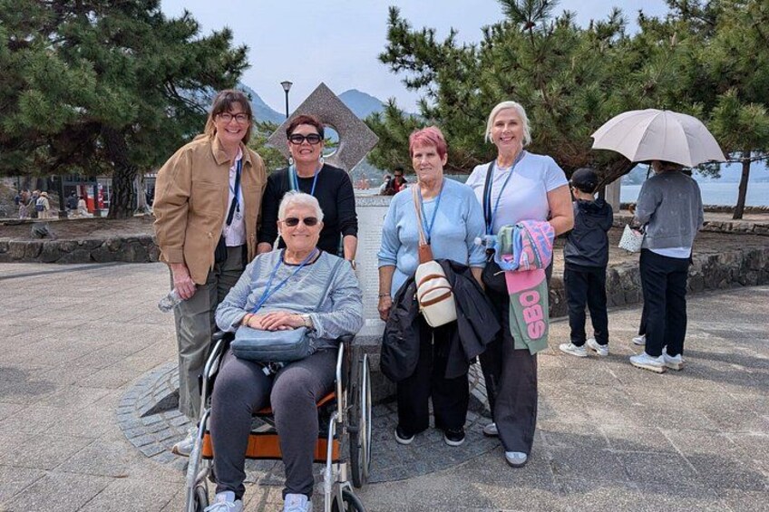 A group of people, including one in a wheelchair, standing in a public space with trees and a body of water in the background. 2