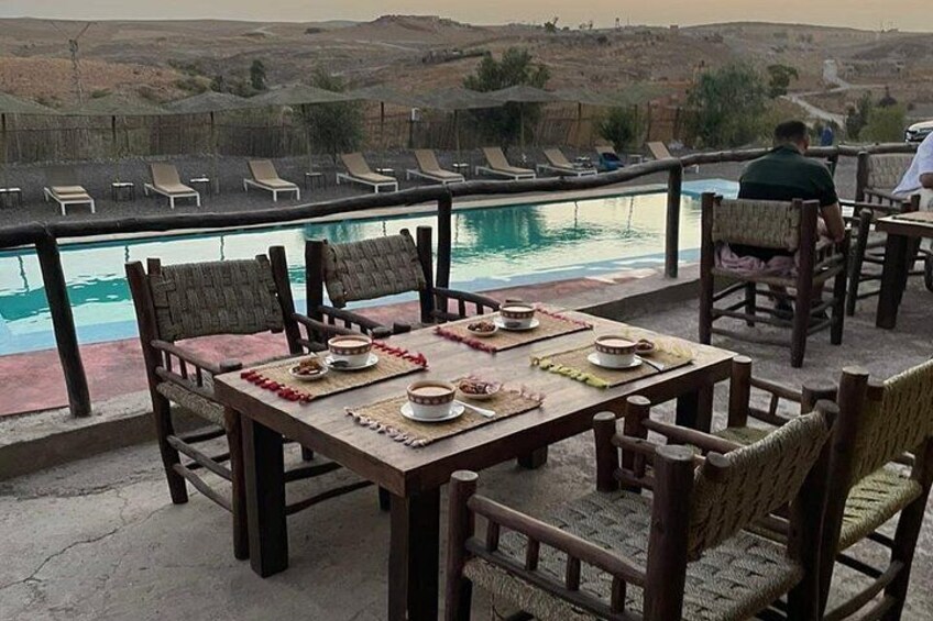 A dining table set for two with chairs, a pool, and a view of hills in the background. 2