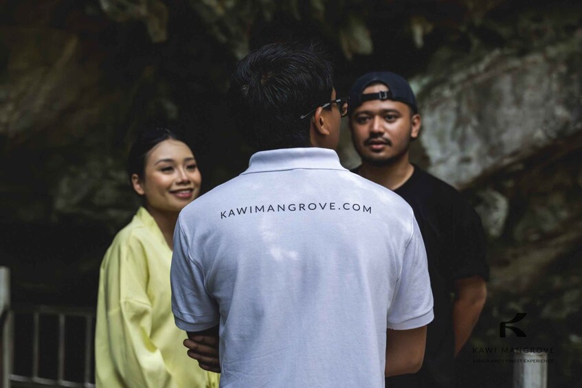 A person in a white shirt with 'KAWIMANGROVE.COM' text, standing with two others in a cave-like setting. 5