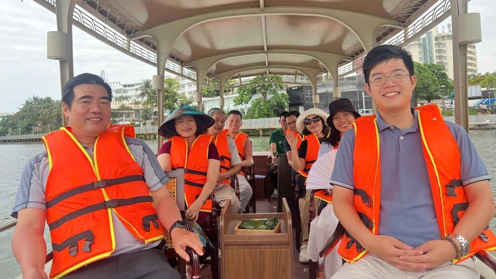 A group of people on a boat, wearing life jackets, with a covered structure overhead and buildings in the background. 5
