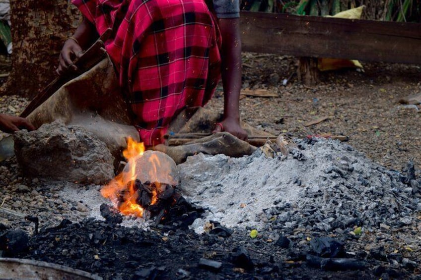 A person tending to a fire with a red and black checkered garment, surrounded by a dirt ground and scattered debris. 4