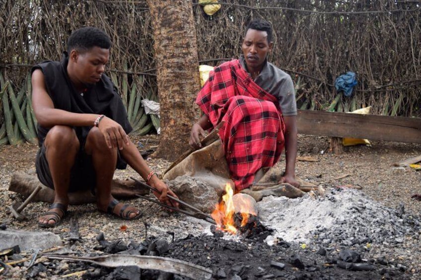 Two people sitting by a campfire in a natural setting with trees and a fence in the background. 2