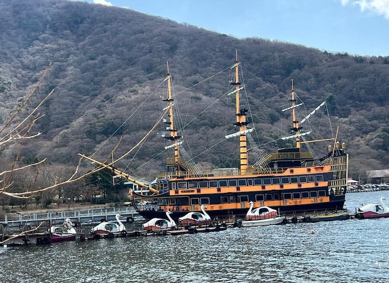 A large orange and black ship docked at a pier with swan-shaped boats in the foreground and a forested hill in the background. 4