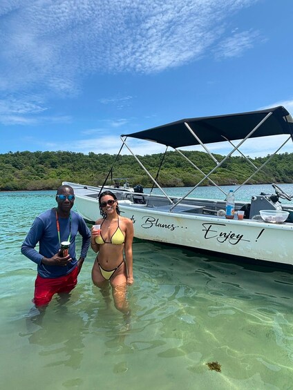 Two people standing in shallow water near a boat with a canopy, with a forested island in the background. 1