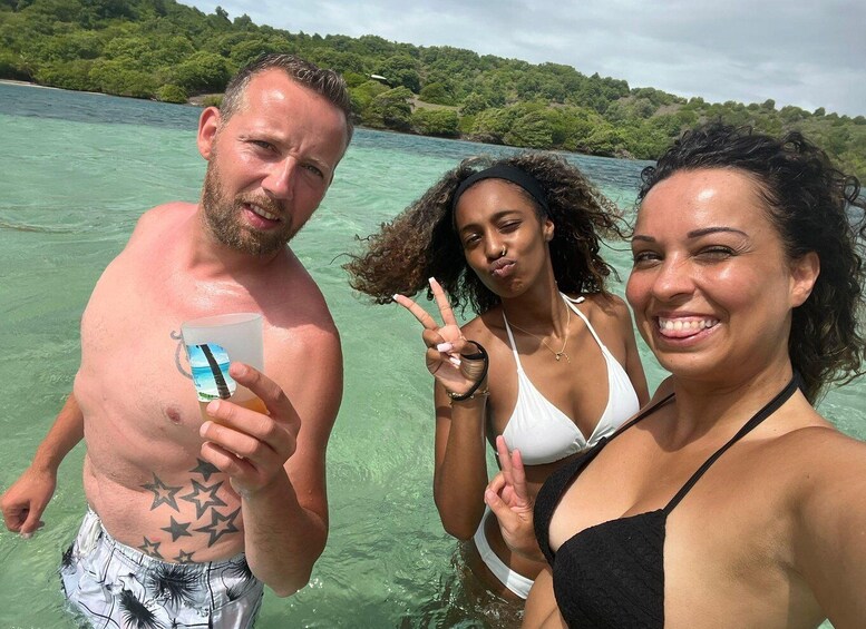 Three people in a tropical beach setting with clear turquoise water and lush greenery in the background. 5