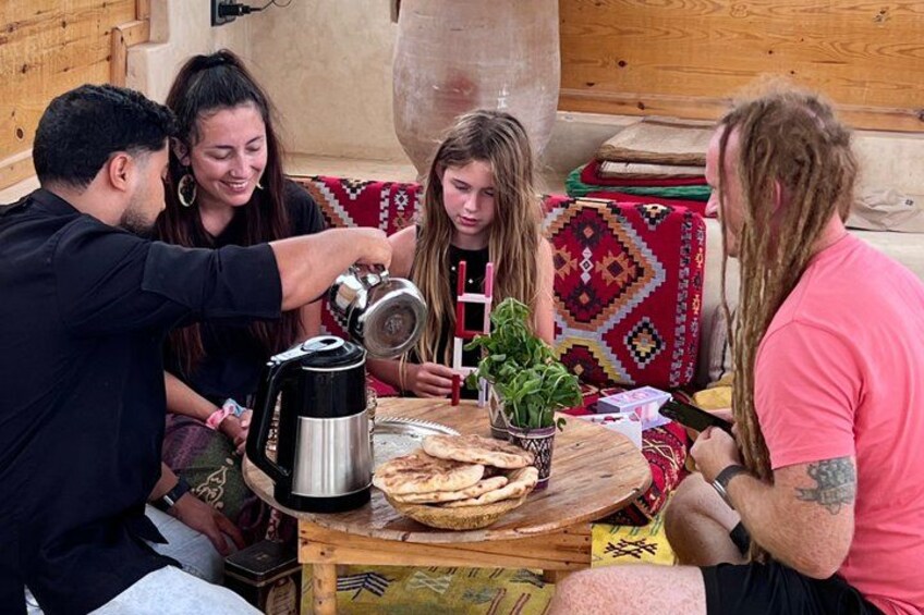 A group of people sitting around a wooden table with a kettle, flatbread, and a potted plant. 1