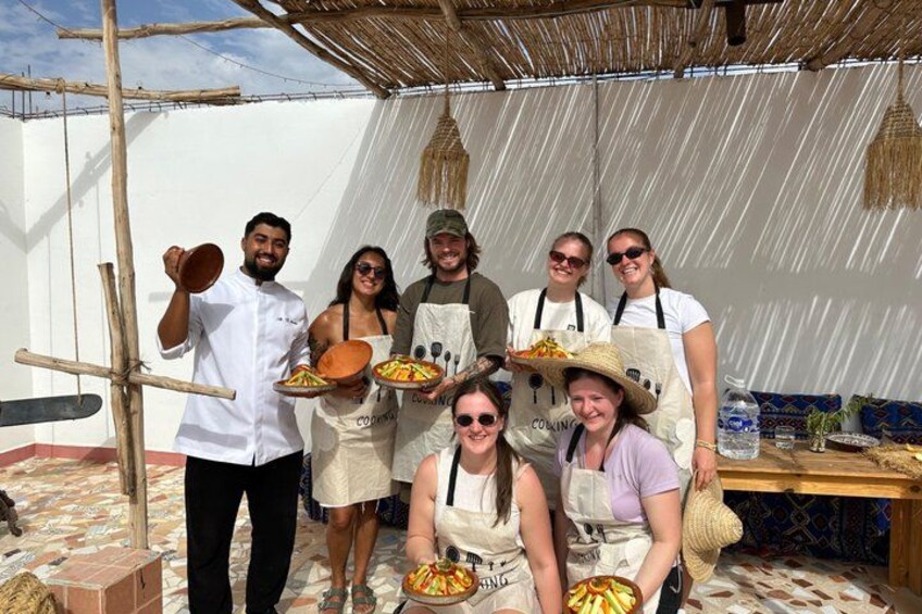 A group of people in a rustic outdoor setting, some holding plates of food, with a thatched roof and a white tent in the background. 3