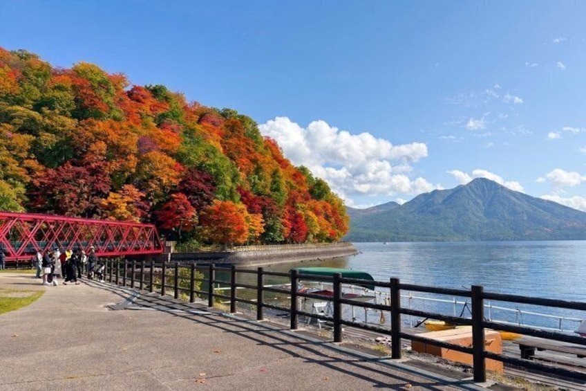 A lakeside walkway with a red bridge, autumn foliage, and a mountain in the background. 2