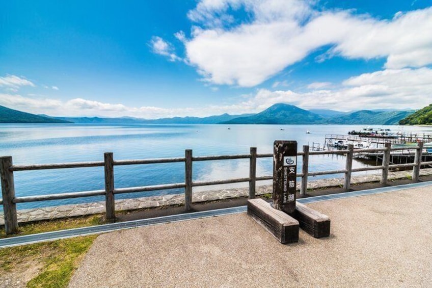 A lakeside walkway with a wooden fence, benches, and a dock with boats. 5