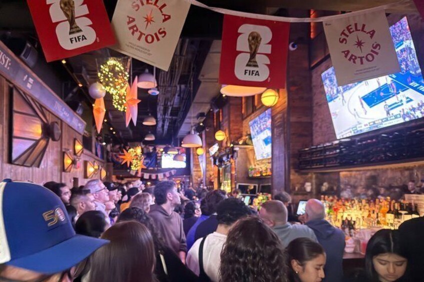 A crowded bar with FIFA and Stella Artois banners, illuminated by hanging lights and decorations. 1