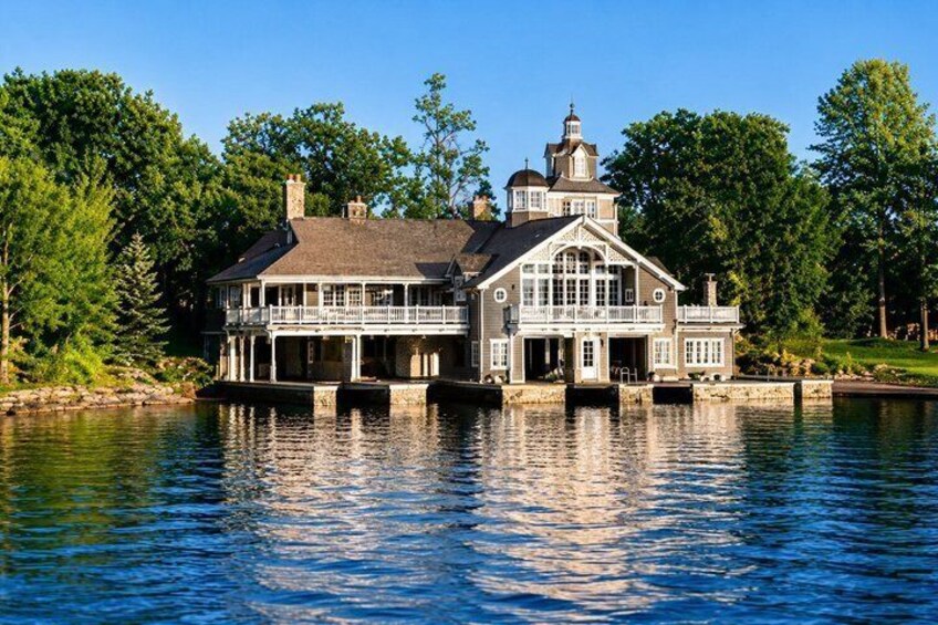 A large, multi-story house with a balcony, situated on a lakeside with trees in the background. 4