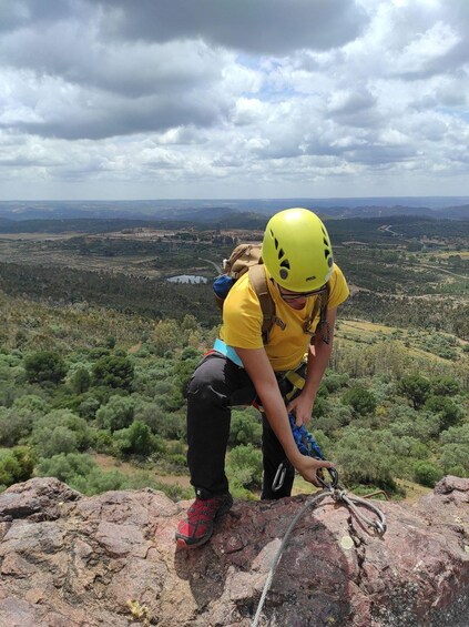 A person in a yellow helmet and yellow shirt climbing a rocky outcrop with a rope, overlooking a valley with trees and distant buildings. 2