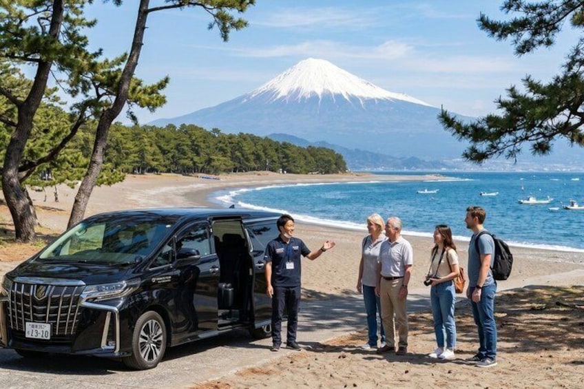 A black van parked on a beach with a group of people standing beside it, a clear view of a snow-capped mountain in the background, and the ocean with boats. 1