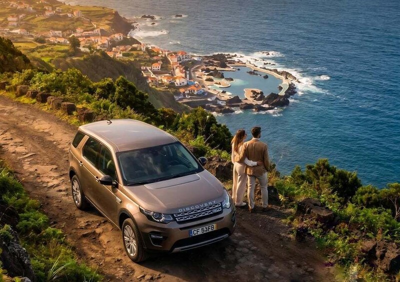 A brown SUV parked on a dirt path overlooking a coastal town with a rocky shoreline and a small bay. 1