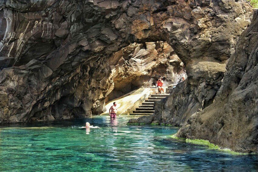 A natural rock arch with clear water, a person swimming, and another person standing on a rock ledge. 5