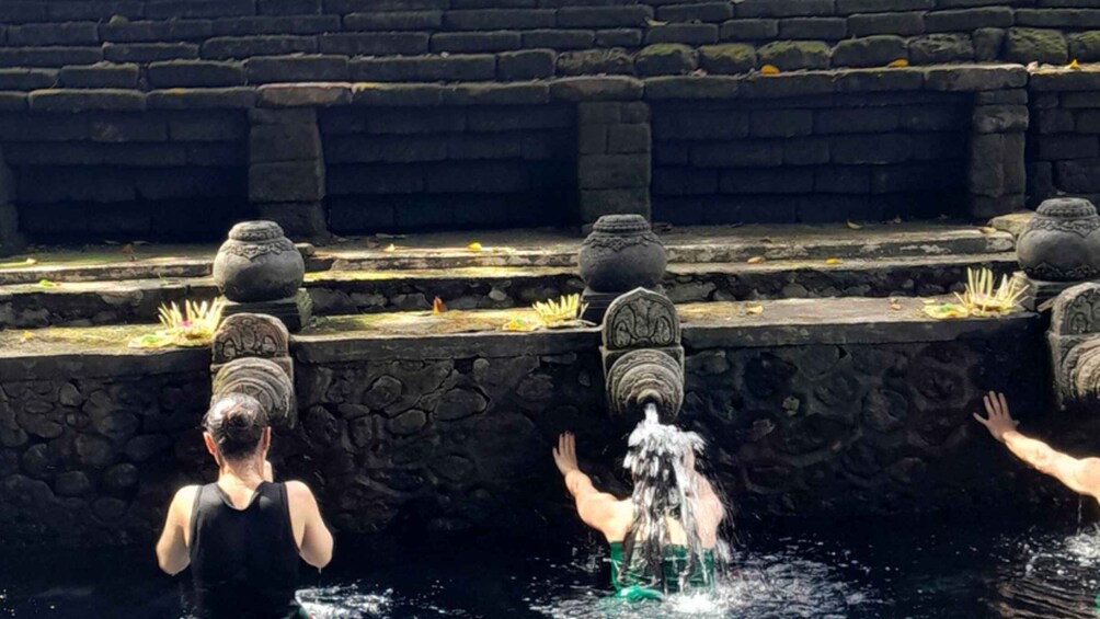 A person in a black tank top standing at a stone water feature with a waterfall, surrounded by stone steps and decorative urns. 1